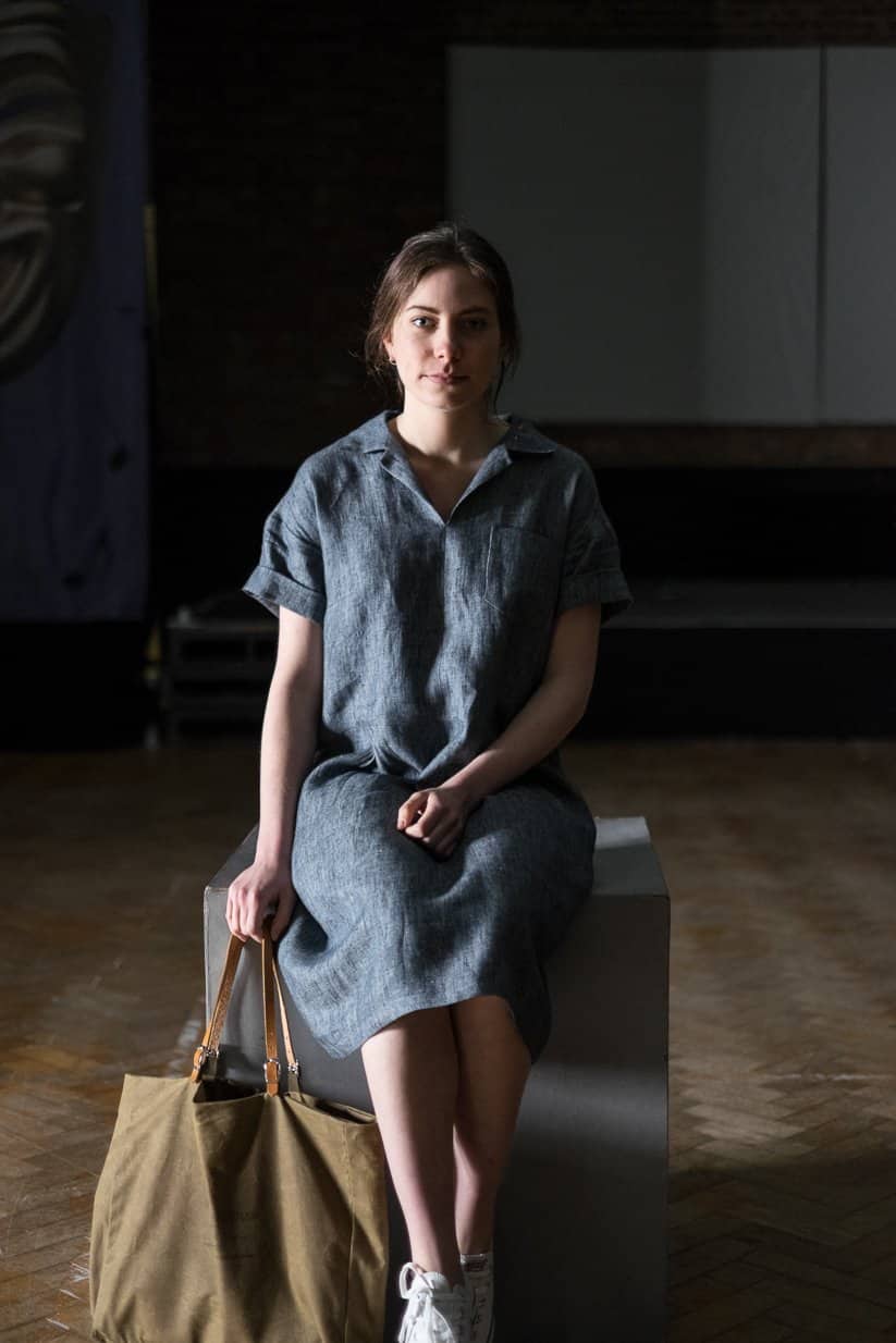 lady sitting. in a dark studio wearing a merchant and mills denim dress in blue